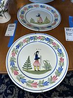 Two Quimper hand-painted plates side by side on a wooden surface, showing traditional Breton figures and floral patterns with metal hangers on back.