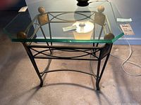 Side table with clear glass top supported by black metal base decorated with four golden brown round accents