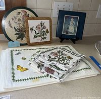 Set of table linens and trivets on kitchen countertop showing round floral tray, square trivet, and stack of printed cloth linens.