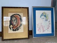Two framed portrait prints displayed side by side, one with a red hood and the other a pastel-colored woman, both signed and numbered.