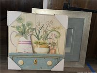 Close-up of the still life framed picture showing potted plants on a blue drawer with white knobs and keyhole, frame has protective corner covers.