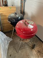 Two dome charcoal grills in a workshop, one red and one black, both with wooden handles and wheels for mobility