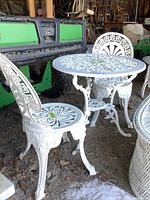 White metal round bistro table and one chair showing detailed cut-out patterns and scrolled legs, placed in a shed setting on a dirt floor.