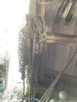 Photo showing the rusty heavy-duty metal chain linked to a wooden block with attached metal hooks, hanging against a wooden wall structure.