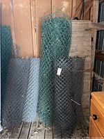 Five rolls of fencing: three silver galvanized chain link fencing, one green vinyl-coated chain link fencing, one black plastic snow fencing rolled and stored vertically in a shed.