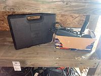 Mastercraft angle grinder with black carrying case and Nail Master electric brad nailer inside cardboard box on wooden shelf.
