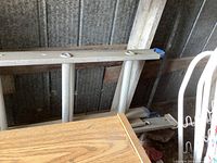 Two aluminum extension ladders leaning against a shed wall with a wooden beam background, showing signs of use with some dirt and dust on the rungs and rails.