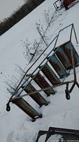 Overall side view of metal rolling ladder showing six perforated steel treads, tubular frame, caster wheels, surface rust, snow background