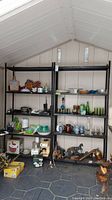 Two black metal shelving units inside a shed, with various household items on shelves including glassware, ceramics, and decorative objects. Focus on shelving unit condition showing paint chips and rust.