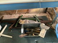 Wide shot showing large vintage hand saws with green frames and wooden handles, placed on the floor behind a plastic basket containing assorted hand tools.