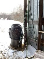 Metal conical trellis and plastic composting bin placed outside near a shed with snow on the ground, showing weathered condition.