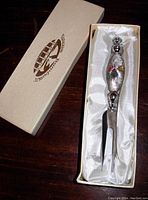 Cheese fork displayed in original box showing the Murano glass handle with millefiori accent and two-prong metal fork end, with satin fabric lining.