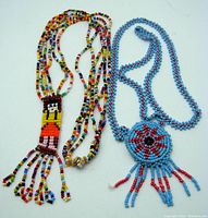 Two vintage indigenous artisan seed bead necklaces laid out, showing one with a multicolored figure pendant and fringe, and the other with a blue circular floral pendant and fringe.