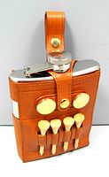 Brown leather golf flask carrier with silver stainless steel flask inside, carrying four wooden tees on front and two circular gold ball markers attached.