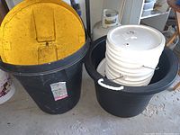 Black 17 gallon garbage can with yellow lid and large black bucket containing a stack of white buckets with lids.