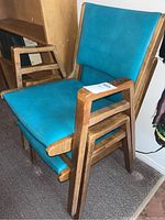Side view of two stackable chairs with wood frames and teal faux leather upholstery showing condition and style.