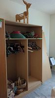 Wide view showing top shelf of wooden shelving unit holding wooden reindeer decoration with red nose, plastic tree stand, and two Christmas wreaths. Lower sections populated with assorted clothing hangers and wicker baskets with gift bags.