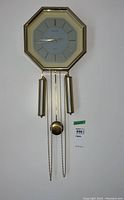 Front view of gold-tone octagonal pendulum wall clock with two weights, pendulum, and chains.