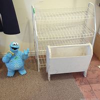 Small white wire shoe rack with three tiers, white wooden magazine rack in front, and blue Cookie Monster figurine next to them.