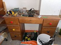 Front view of wooden seven drawer desk with colorful knobs, cluttered surface, and items below it. Shows need for cleaning and signs of rodent evidence.
