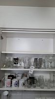Two shelves in a white cabinet filled with assorted clear glasses and ceramic mugs with different designs.