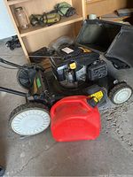 Side view of black Yard-Man push lawn mower with attached grass catcher bag and red plastic jerry can in front.