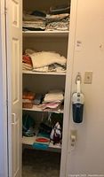 View of linen closet with folded linens on three shelves, dustbuster vacuum hanging on wall, small iron and sewing items on lower shelf.