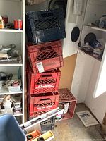 Stack of 6 plastic milk crates in red and black/dark blue colors in a storage area with surrounding tools and materials.