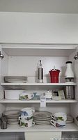Full view of dishware cabinet shelves containing plates, illustrated bowls and cups, casserole dish, kitchen chopper, and oil dispenser