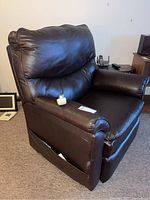 Brown leather reclining lift chair upright with remote on seat. Shows minor wear and two torn spots near side pocket.