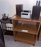 Wooden shelf with stereo system on top, and smaller round side table with telephone and notepad on top, visible damage on side table top
