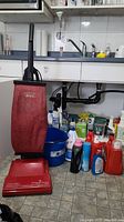 Hoover Encore upright vacuum cleaner with red bag standing on tiled floor next to multiple plastic bottles of cleaning and laundry supplies arranged under a sink area.