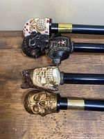 Four walking canes laying flat showing detailed decorative skull handles and black shafts with metal bands.
