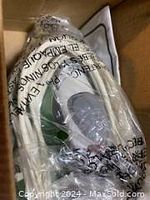 Power cord wrapped in plastic packaging inside box, showing the cord and plastic wrap.