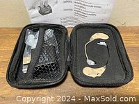 Two beige personal sound amplifiers placed inside a black zippered carrying case, with compartments holding accessories and the user manual in the background.