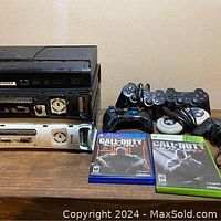 Photo shows three stacked video game consoles including a black Xbox 360, silver and black PlayStation consoles, several controllers and two video games Call of Duty Black Ops II and Call of Duty Black Ops.