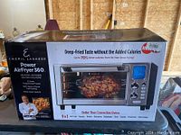 Box of Emeril Lagasse Power AirFryer 360 air fryer oven, showing front with marketing text and image of rotisserie chicken inside appliance.