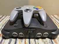 Black Nintendo 64 console with gray Nintendo 64 controller on top, showing front logos and controller buttons.