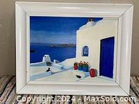 Front view of the framed painting showing white architecture, blue door and window, red and blue flower pots, and a black cat with sea view behind.