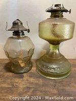 Photo showing two vintage oil lamps side by side, one green textured glass with a pedestal base, the other clear faceted glass.