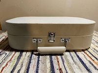 Closed view of white textured suitcase-style portable record player with handle and metal clasps on carpet.