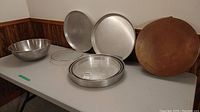 Full view of the bakeware items displayed on table: stainless steel bowl, three round cake pans, cooling rack, and copper finish cake pan with wear and dents.