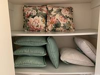 Four solid green cushions stacked in a pile with two white cushions placed flat next to them, all on a white shelf.