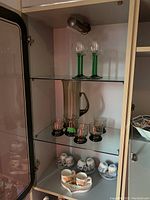 Cabinet shelf with four green-stemmed wine glasses on top, glass jug and five small glasses on the middle shelf, and four sets of small coffee cups with saucers on the bottom shelf along with a white ceramic tray.