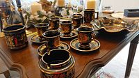 Demi tasse cups and saucers, mugs, and decorative bowls featuring 24K gold leaf Greek designs arranged on a wooden table.