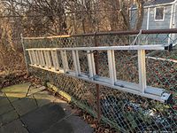 Photo of a silver metal ladder hanging on a chain link fence outdoors on a paved surface with dead leaves around.