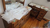 Various linens and tablecloths stacked on a wooden dresser and chair with some pieces covered in plastic for protection.