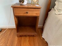 Front view of wood night table showing single drawer and open bottom shelf, positioned next to bed with wood floor underneath.