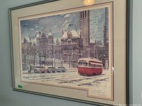 Framed signed print by B. Liaskas showing an urban snowy street scene with a red streetcar and historic buildings, in a silver frame hung on a wall.