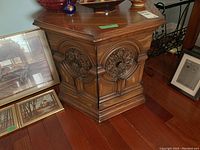 Wooden hexagonal side table with carved medallion detailing and contents removed, shown from an angle highlighting the front and side. Door is off hinge, showing damage and wear on base and door edges.